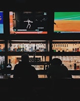 A bar counter with several people sitting in front of it is shown. Above the counter, there are multiple television screens displaying sports broadcasts, including football and baseball. Behind the counter, various bottles of alcohol are neatly arranged.