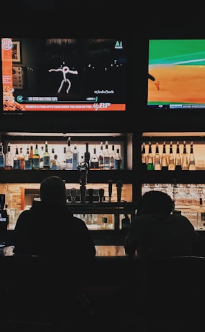 A bar counter with several people sitting in front of it is shown. Above the counter, there are multiple television screens displaying sports broadcasts, including football and baseball. Behind the counter, various bottles of alcohol are neatly arranged.