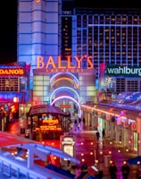 A brightly lit urban scene featuring the entrance to Bally's Las Vegas, surrounded by various illuminated shop signs including Giordano's, Wahlburgers, and Redneck Riviera. The street is bustling with people and rich with neon lights and colorful signage, creating a lively nighttime atmosphere.