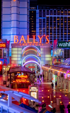 A brightly lit urban scene featuring the entrance to Bally's Las Vegas, surrounded by various illuminated shop signs including Giordano's, Wahlburgers, and Redneck Riviera. The street is bustling with people and rich with neon lights and colorful signage, creating a lively nighttime atmosphere.
