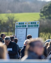 A group of people gathered outdoors with two large betting boards in the background labeled 'REG DAVIES'. The setting appears to be a countryside location with trees and fields visible in the distance. The crowd consists of individuals dressed in jackets and hats, engaging in conversation.