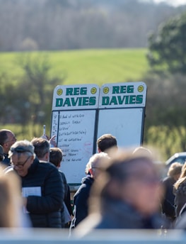 A group of people gathered outdoors with two large betting boards in the background labeled 'REG DAVIES'. The setting appears to be a countryside location with trees and fields visible in the distance. The crowd consists of individuals dressed in jackets and hats, engaging in conversation.