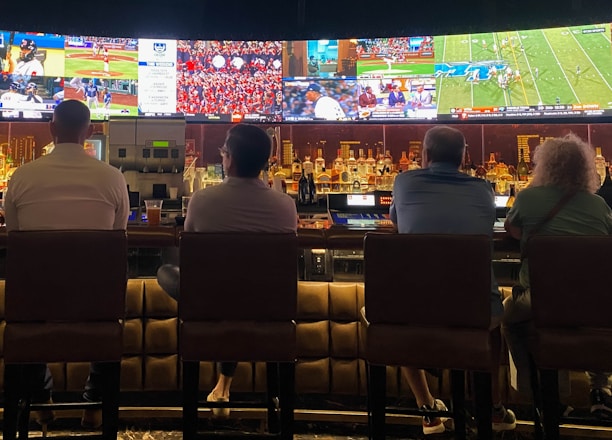A group of people sit at a bar with their backs to the camera, watching multiple sports games displayed on large screens. The bar counter is lined with various bottles of liquor, and the atmosphere suggests a casual and relaxed social setting.
