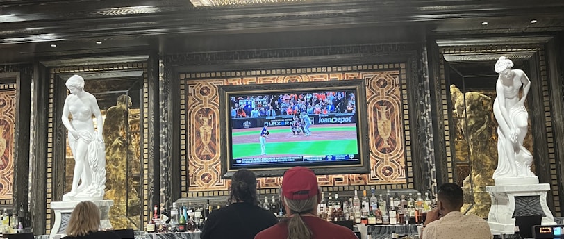 Several people are seated at a luxurious bar, with a large TV screen showing a sports game. The bar is elegantly decorated with ornate details and sculptures. The ceiling has a large, rectangular, crystal chandelier.