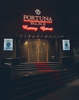 A neon-lit casino entrance with a red carpet leading up to large double doors. The sign reads 'Fortuna Palace Luxury Games' in bright red and white neon lights. The dimly lit atmosphere suggests a night-time setting. There are two digital displays on either side of the entrance showing promotional content.