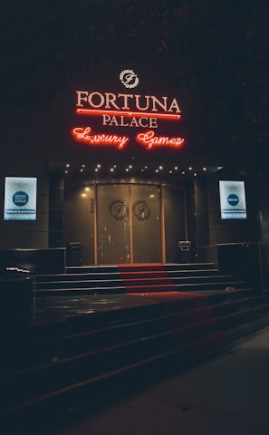 A neon-lit casino entrance with a red carpet leading up to large double doors. The sign reads 'Fortuna Palace Luxury Games' in bright red and white neon lights. The dimly lit atmosphere suggests a night-time setting. There are two digital displays on either side of the entrance showing promotional content.