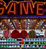 A brightly lit carnival game stall with a large 'GAMES' sign at the top surrounded by red and yellow lights. Various sports balls, including basketballs and soccer balls, are hanging from the ceiling and neatly arranged on shelves below.