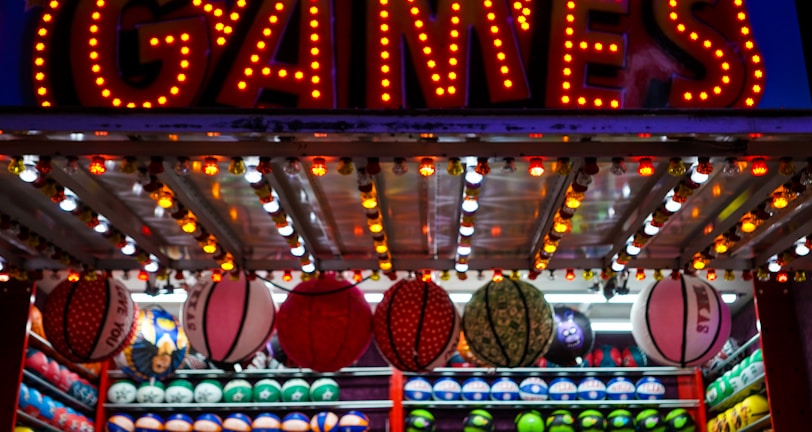 A brightly lit carnival game stall with a large 'GAMES' sign at the top surrounded by red and yellow lights. Various sports balls, including basketballs and soccer balls, are hanging from the ceiling and neatly arranged on shelves below.